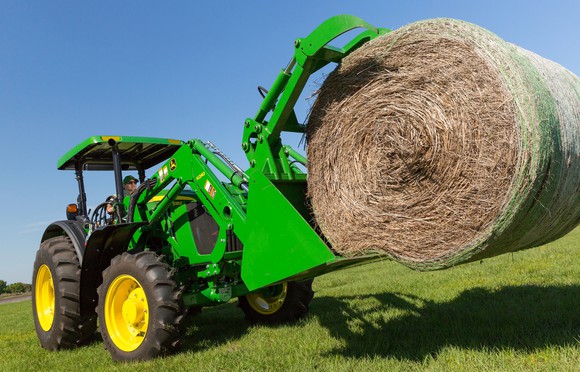 Green Deere farming equipment picking up a hay bale in an open field.