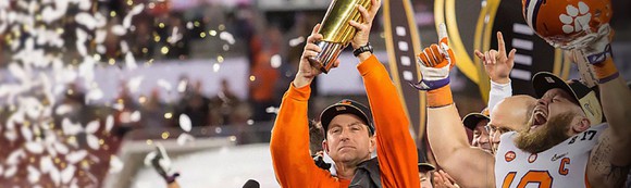 Clemson coach holding up national championship trophy with team celebrating.