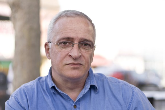 An older man with glasses and a blue button-down shirt sits outside with a serious expression.