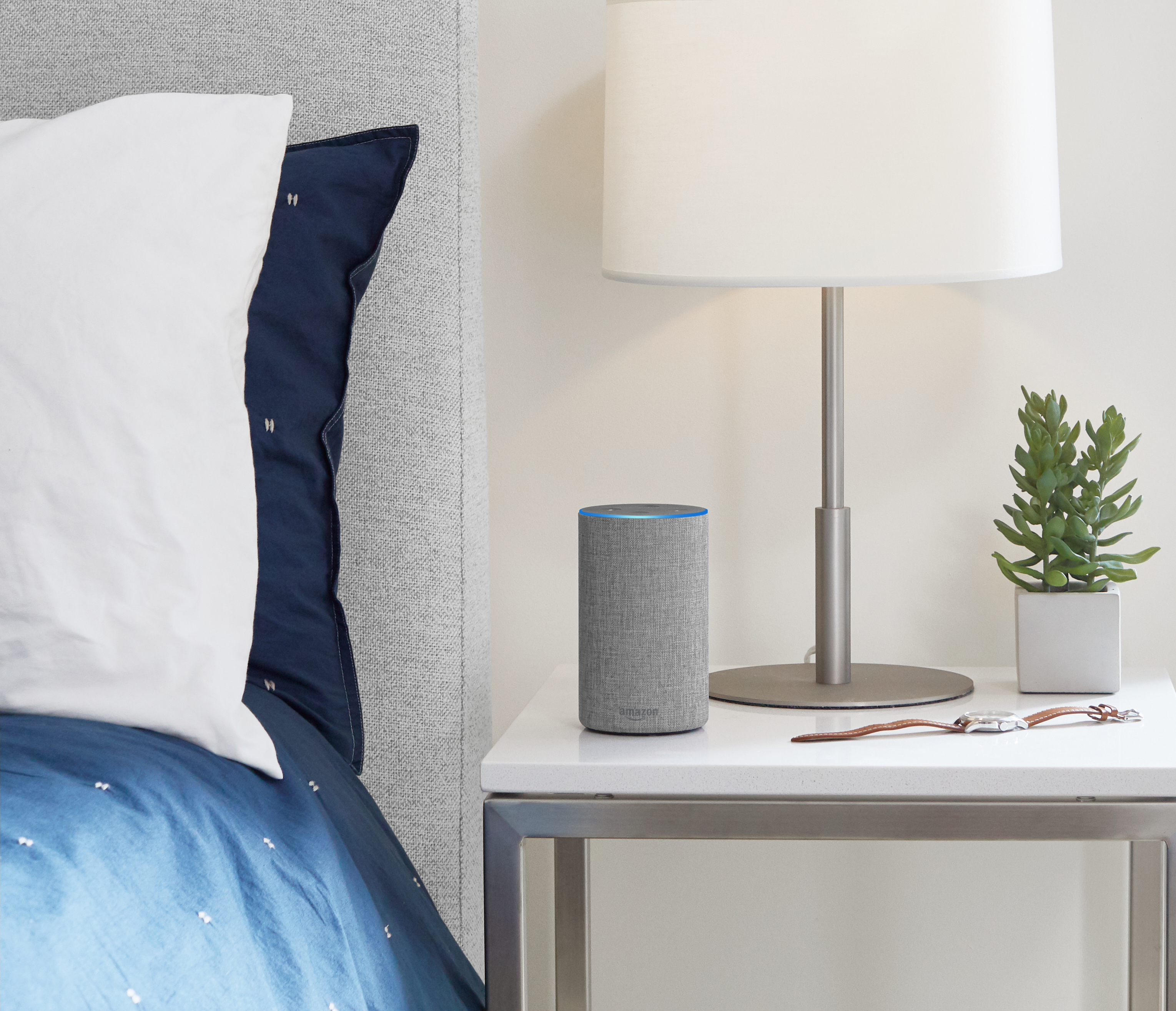 A gray Amazon Echo smart speaker on a nightstand next to a bed.
