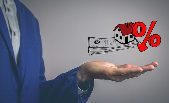 man in business suit holding his hand below an illustration of a house and a percentage symbol sitting on top of a stack of cash