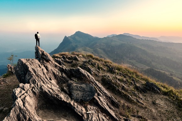 A person stands on top of a mountain staring at a mountain range in the distance.