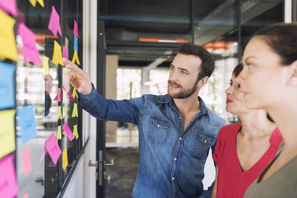 Three businesspeople in casual clothes talking in front of a wall filled with post-its.