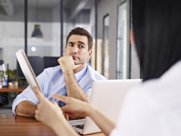 Skeptical looking man glares across a table at someone holding a notebook and pointing to it.