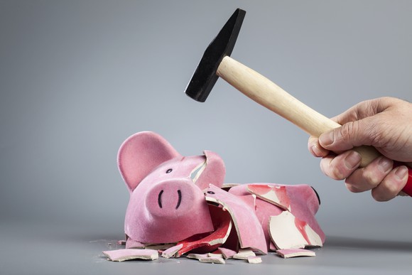 A man's hand holds a hammer above a broken piggy bank