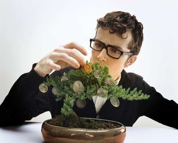 A young, bespectacled man tending a bonsai tree with coins hanging on the branches