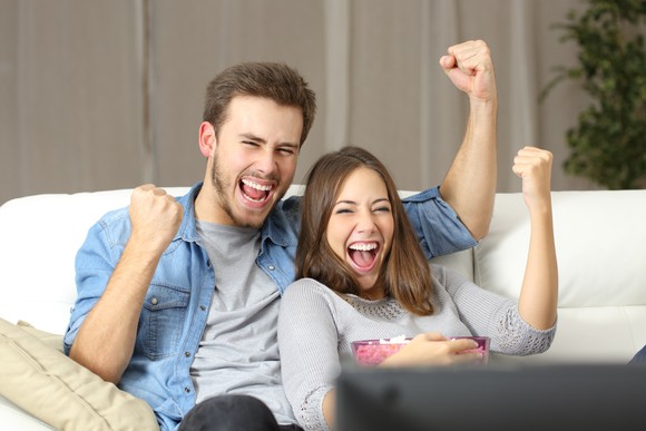 A young, happy couple watching TV, smiling, and pumping their fists in excitement.