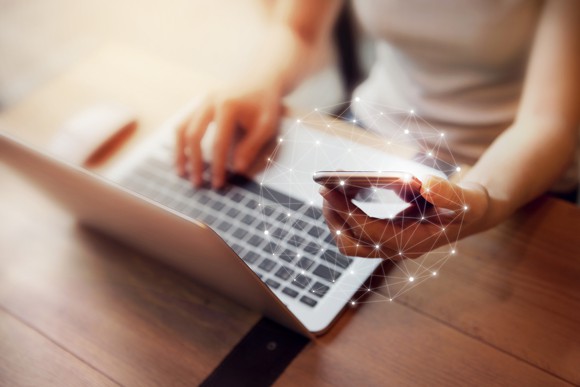 Woman types on laptop while holding a cell phone in her hand with a glowing globe surrounding it.