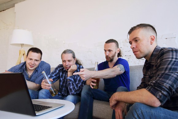 Friends sit on a sofa as they watch a laptop computer on the table.