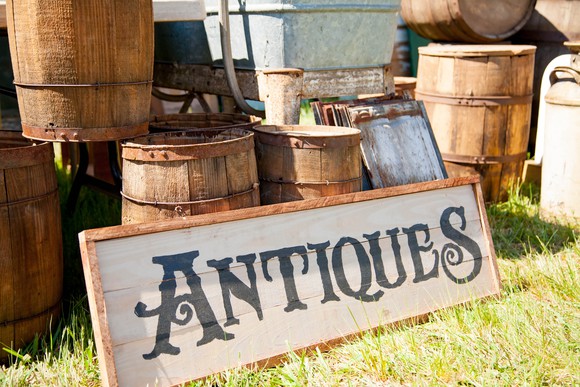 Sign with the word antiques on it, resting outdoors on the grass against a pile of wooden barrels.