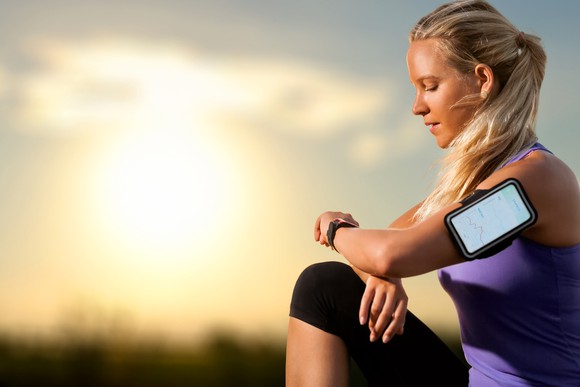A blonde-haired woman in a purple shirt and black leggings takes a break from a run to check her wearable fitness device at sunset