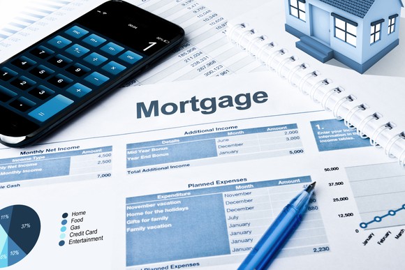 Calculator, toy house, and pen on top of documents labeled mortgage with graphs, charts, and calculations.