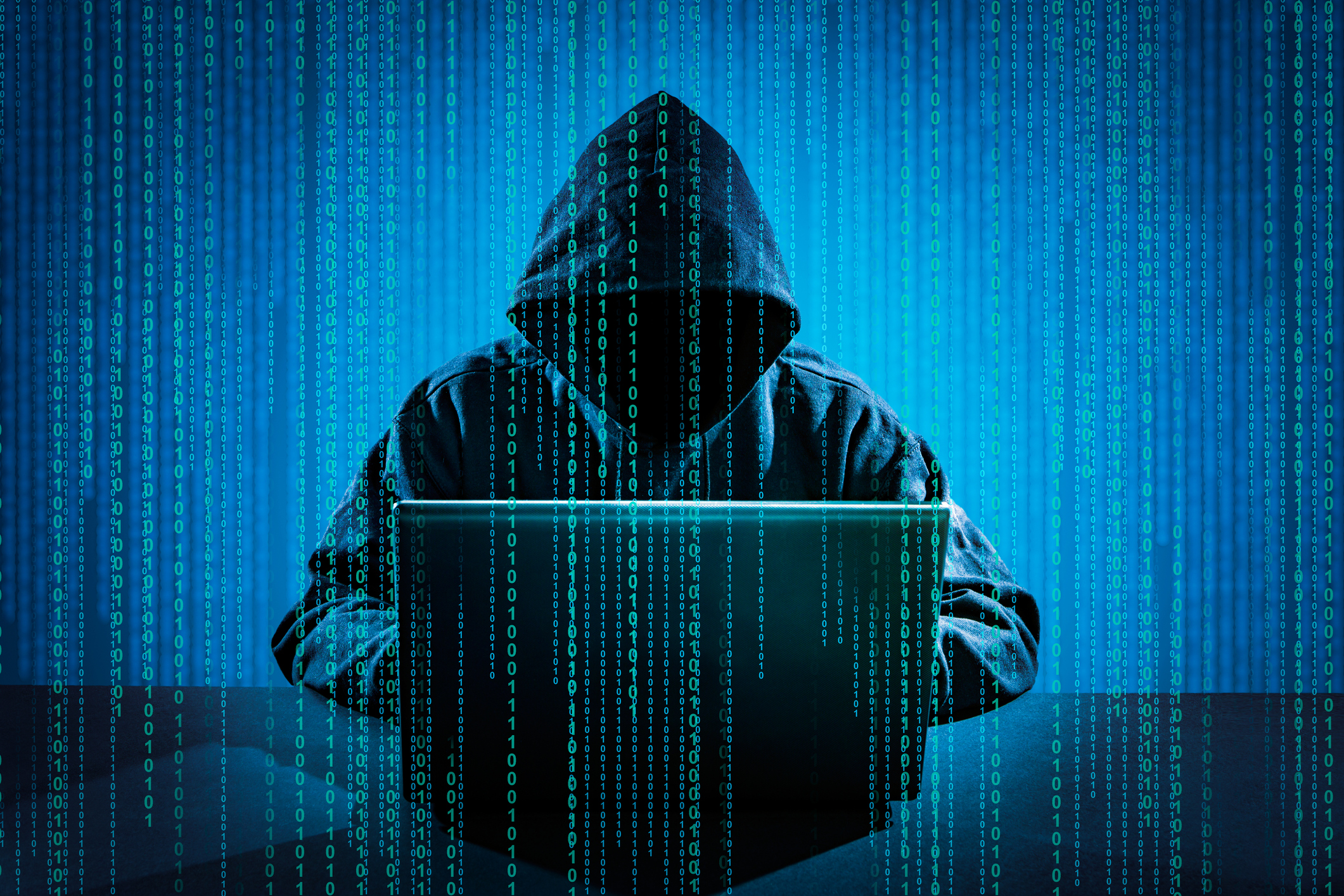 A person shrouded in a dark hood sitting in front of a computer