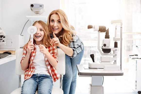 A woman standing behind a girl who is getting her eyesight checked in an office.