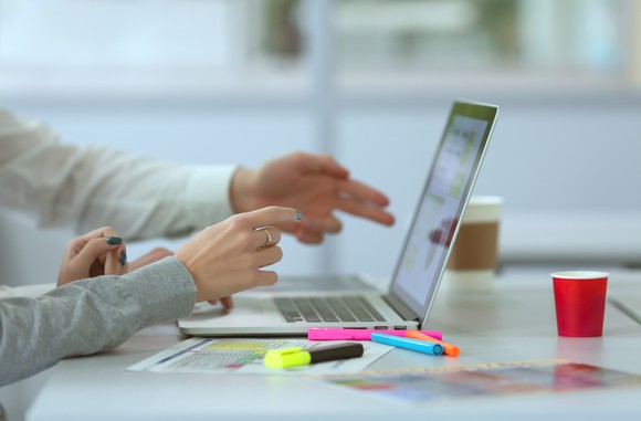 Hands pointed toward laptop screen with papers and highlighters next to it