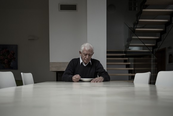 elderly man sitting alone at large table in gray house and eating from a bowl