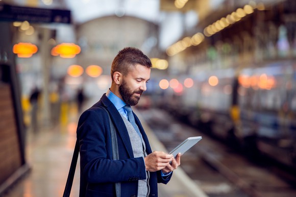 A smiling man in a suit stands on a platform in a train station, looking down at a tablet in his hands.