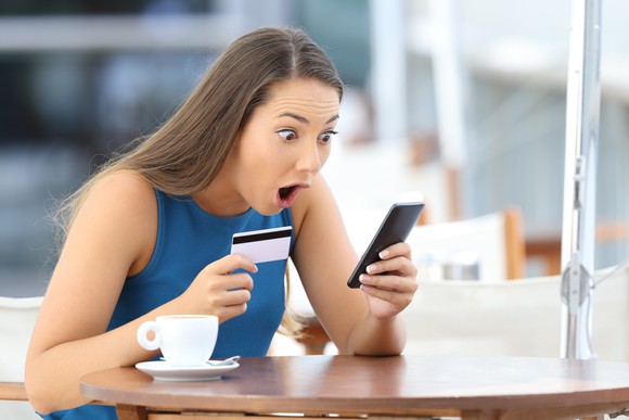 young woman holding credit card in one hand while looking at smartphone with an astonished expression