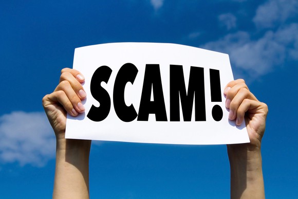 two hands holding up a sign that says scam, with an exclamation mark