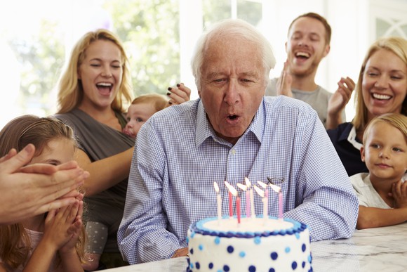 Senior male blowing out candles on a birthday cake with people of various ages clapping behind him