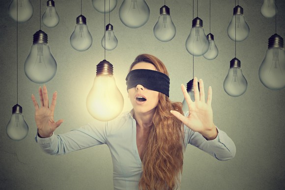 A blindfolded woman makes her way among hanging lightbulbs, seeking the bright one