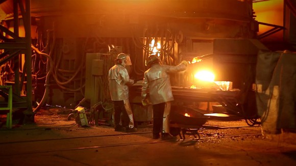 Two workers in heat-protection suits doing metal work in front of a glowing furnace.