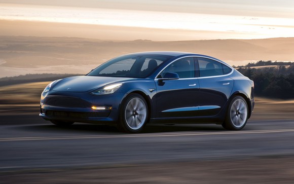 Dark-colored Model 3 sedan on a road with a hilly open landscape beyond.