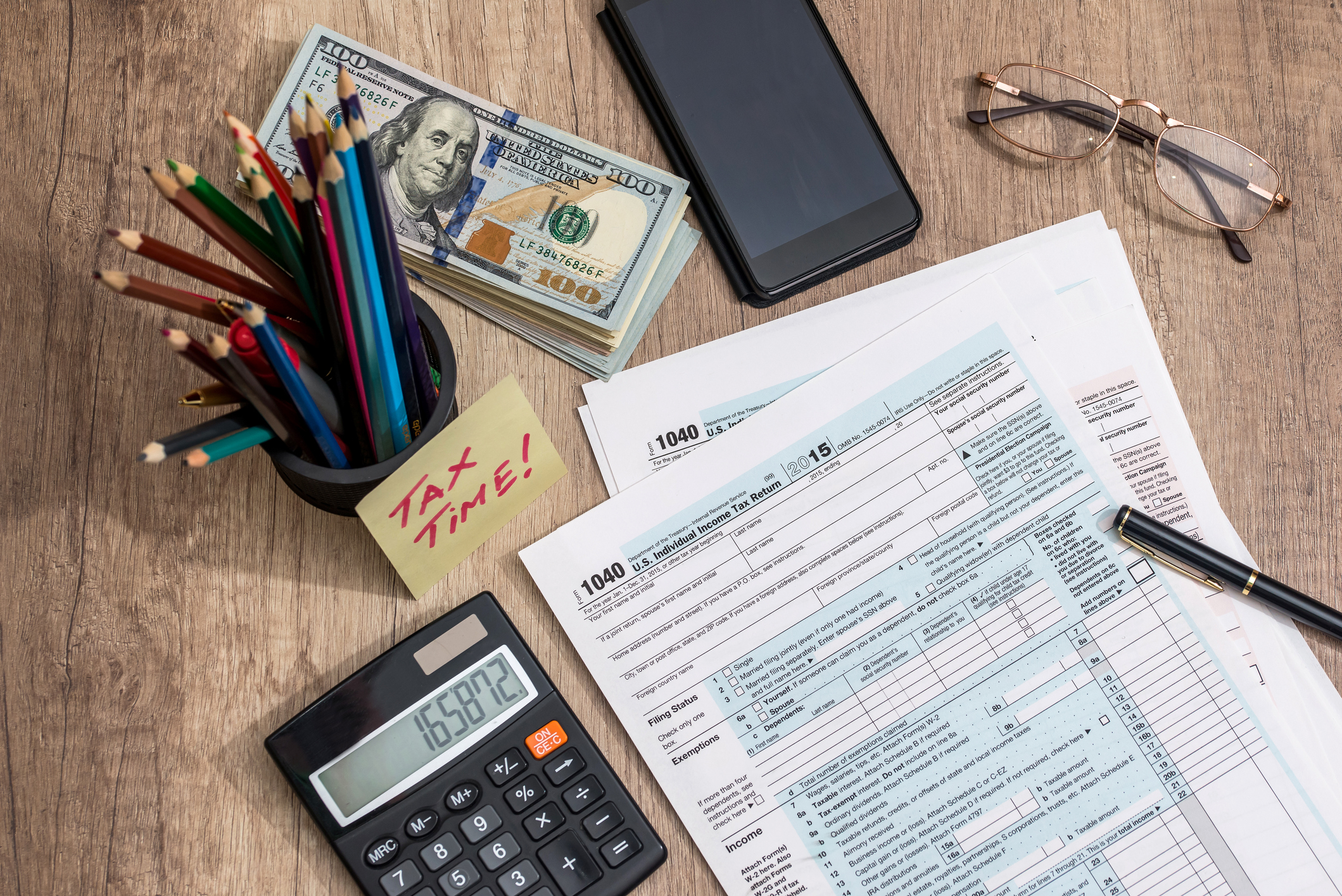Tax forms on table, next to calculator, stack of money, glasses, and cup of pencils.