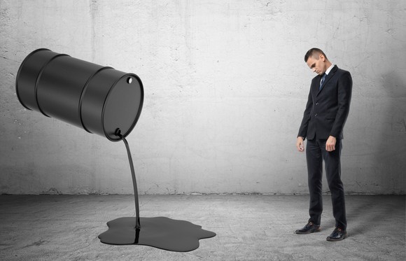 A frowning man stands next to an oil drum spilling oil on the ground.