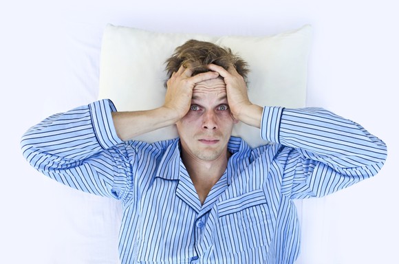 A man in pajamas lying in bed, with his hands to his head and eyes open.