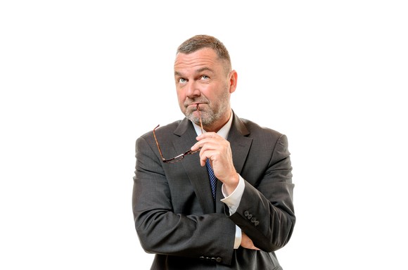 A man in a suit, looking upward, and holding the ear piece of his glasses pensively to his mouth.