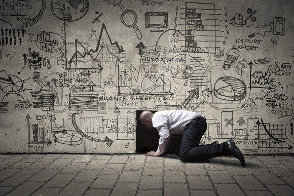 A man on his knees looking through a hole in a wall full of business diagrams