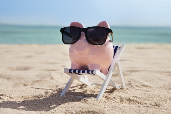 A piggy bank with sunglasses sits on a beach chair on a beach with a body of water in the background.