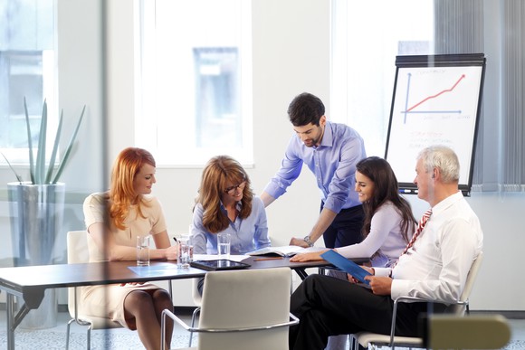 A man in an office setting stands over a group of others around a table, reviewing papers, with a chart on a stand behind them.
