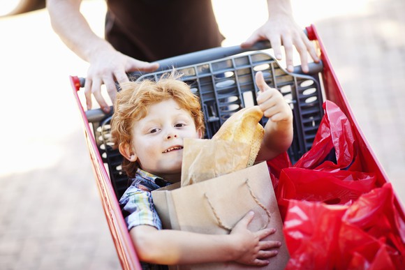 A small boy being pushed in a shopping cart clutches a bag while giving a thumbs-up sign.