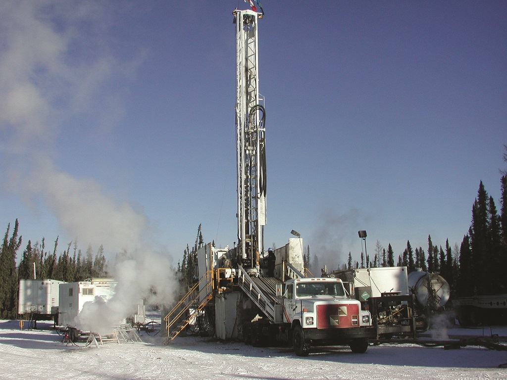 Drilling rig with associated buildings in a winter setting with snow on the ground and pine trees surrounding the area.