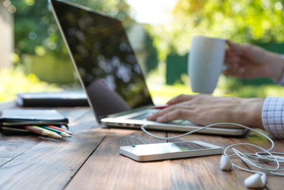 A man sitting in a park, using a laptop, and holding a cup of coffee. On the picnic table are a smartphone and headphones.
