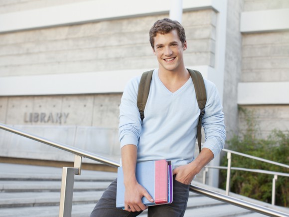 Male student holding books on stair rail outside.