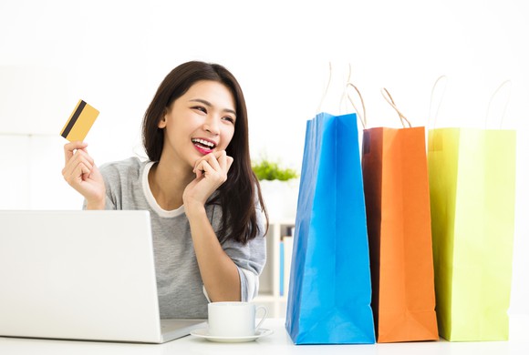 Smiling woman holding a credit card while sitting in front of a laptop and looking at shopping bags on the table.