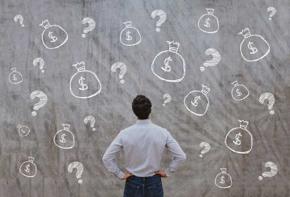 A man standing with his hands on his hips and looking at a chalkboard with bags of money and question marks drawn on it.
