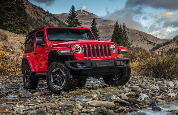 A red 2018 Jeep Wrangler SUV is parked next to a stream near a rough mountain trail.