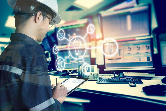 A man wearing a hard hat using a tablet in room of computer monitors. An overlay of industrial tool icons.