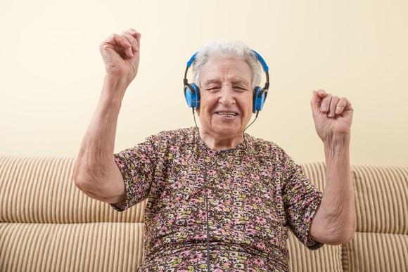 A happy senior woman listening to headphones and raising her arms as if dancing.