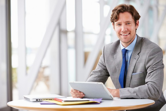 Smiling man in business suit sits at a small round table, holding a tablet, with a laptop and papers spread out in front of him.
