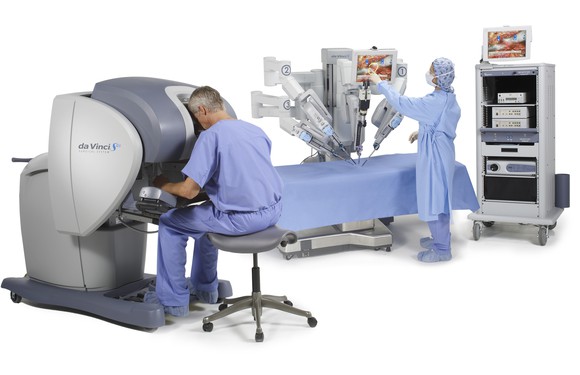 A surgeon seated at a console which a scrubbed nurse adjusts the arms on a surgical robot.