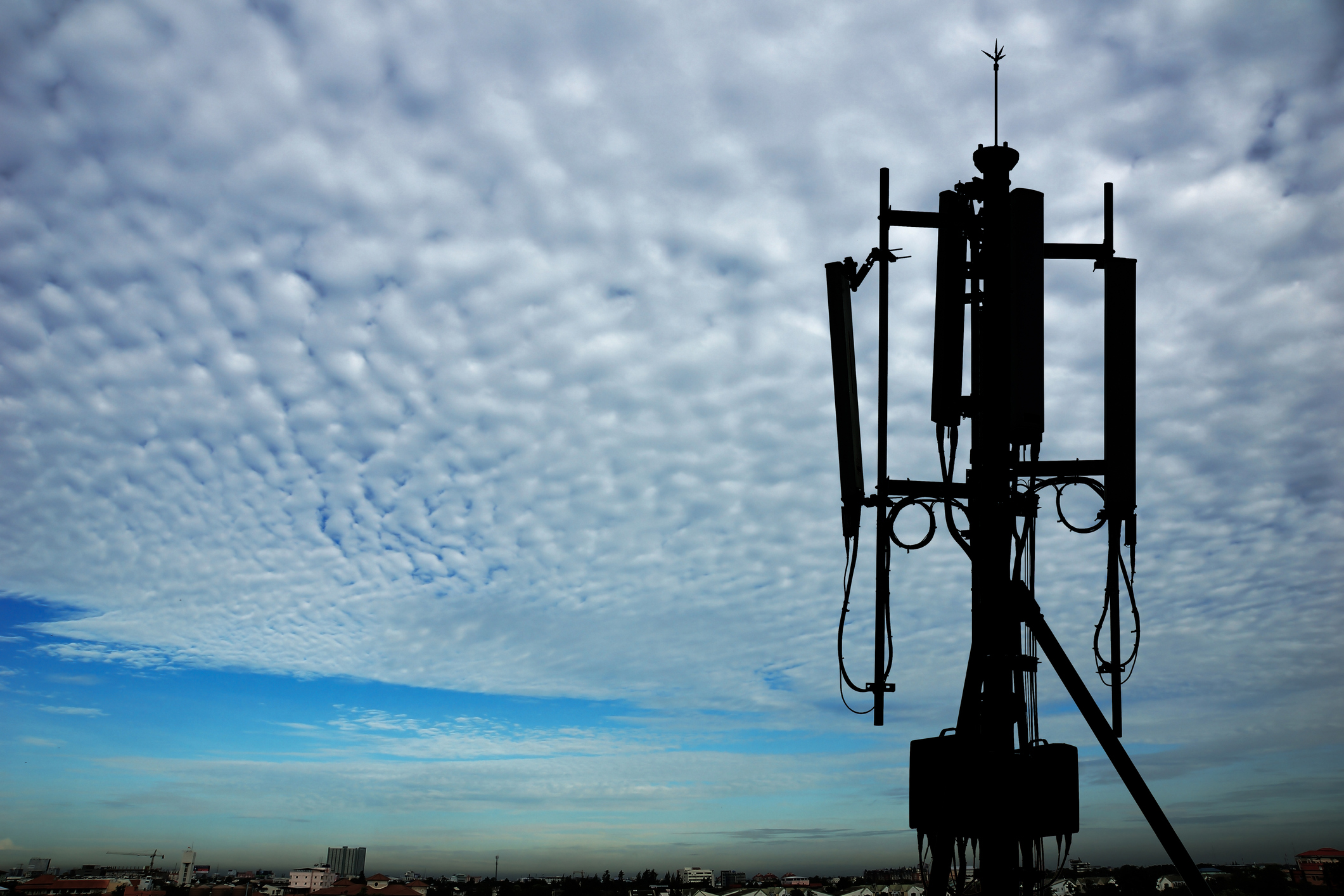 A radio transceiver atop a cell tower against cloudy skies and a faraway city skyline.