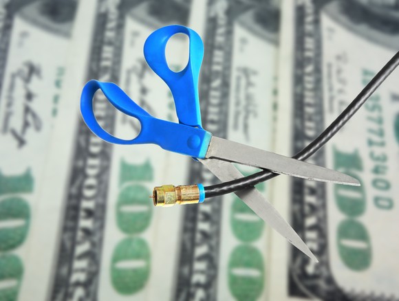 A pair of scissors cuts a cable cord, with one hundred dollar bills in the background.