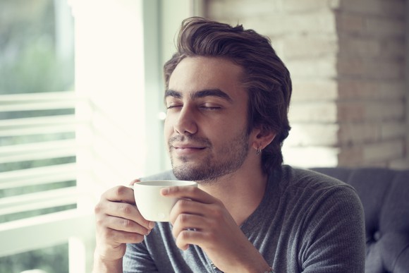 A man, seated by a window, smiles with his eyes closed as he cradles a cup in both hands.