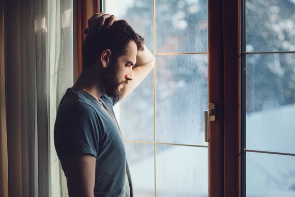 A man stands before a glass door, head on hand, head bowed, and eyes closed.
