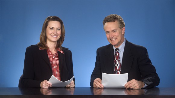 A woman and man, both smiling and dressed in suits, sit behind a desk with papers in hand looking at the camera.